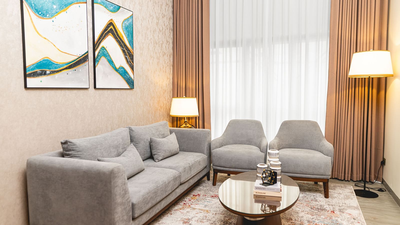Room at KertelSuites with a sofa, two matching armchairs, a round coffee table, two floor lamps, a patterned rug, and abstract art on the wall. Large windows with sheer curtains allow natural light in.