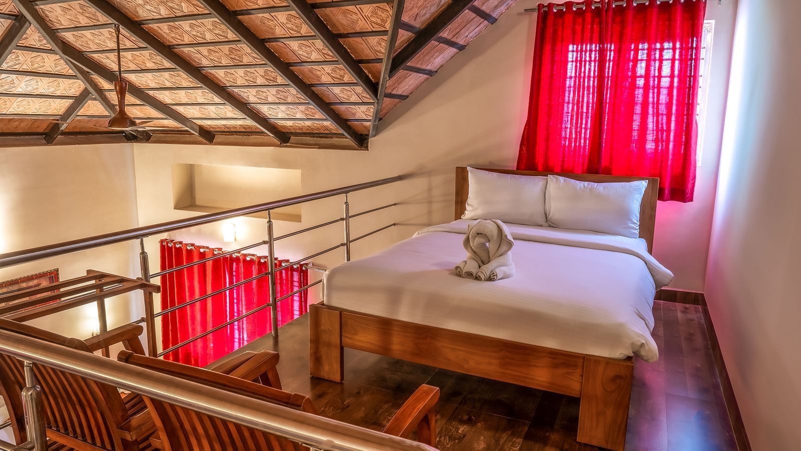 An elevated view shows Attic Villa featuring a wooden bed frame, white linens, and red curtains, with a glimpse of the room's upper level | Woodstock Resorts, Coorg