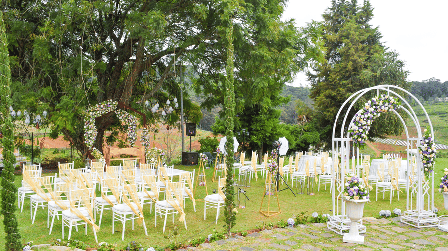 An outdoor wedding or event setup on a green lawn, featuring two white archways decorated with flowers and numerous white chairs with yellow sashes, surrounded by trees.
