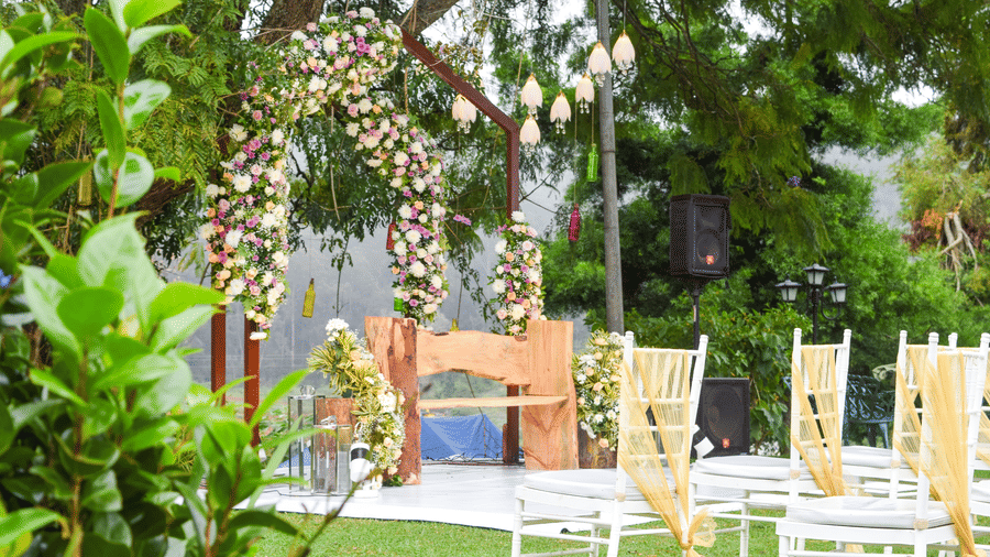 A charming outdoor wedding or event area featuring a rustic wooden archway adorned with white and pink flowers, with white chairs and tables set on a grassy area, surrounded by lush green plants.