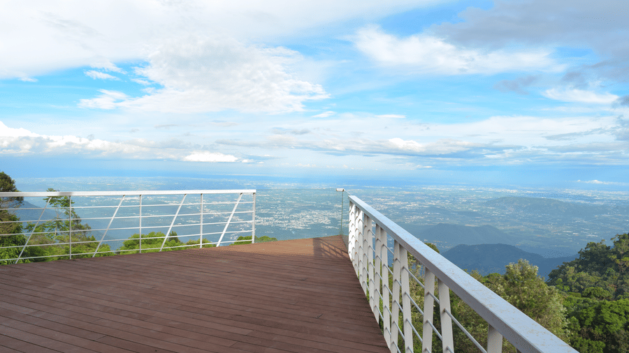 Exterior side view of a white, modern villa with balcony, perched on a green slope with ocean in the distance.