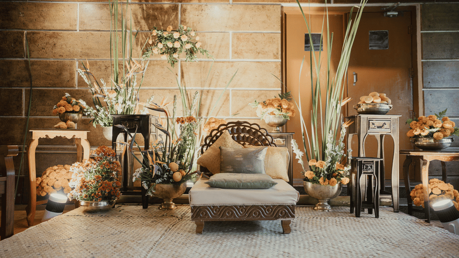 An indoor lounge area with a rustic aesthetic, featuring a light-colored sofa with patterned cushions, surrounded by various floral arrangements, decorative furniture, and tall plants against a textured wall.