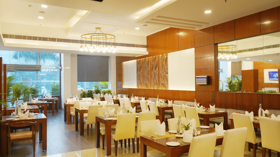 Spacious and elegant dining room at Nemesia with multiple tables set with white tablecloths and chairs. The room features large windows, wooden paneling, and a chandelier, creating a warm and inviting atmosphere.
