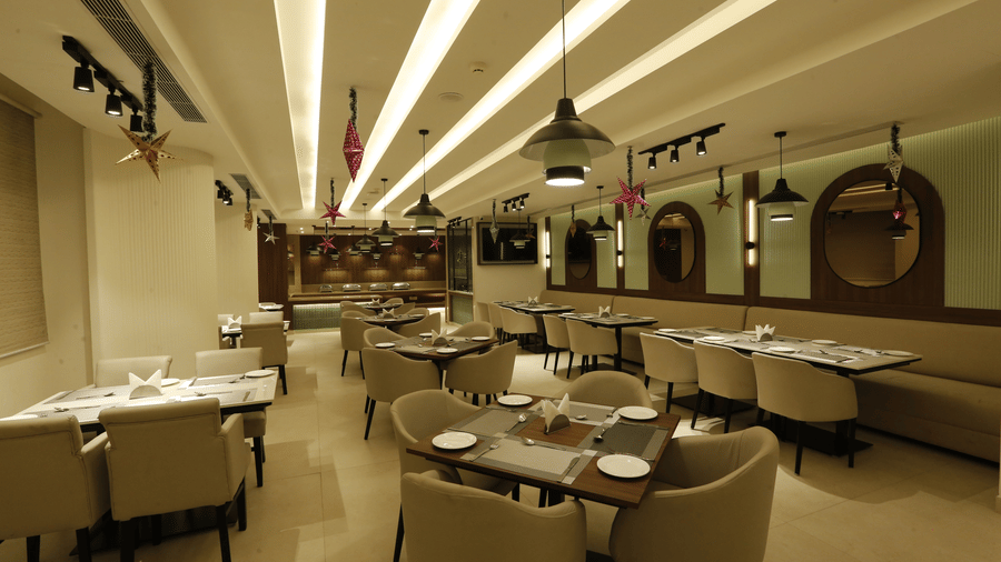  A poster of a restaurant at Tulip Inn, Zirakpur featuring several sets of tables, chairs, and sofa, lights hanging from the ceiling and mirrors attached on the wall