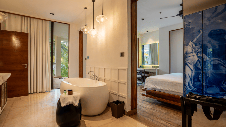  A luxurious hotel room with a white bathtub next to a large window and a view into the bedroom. The room is decorated with blue tiles and reflective surfaces.