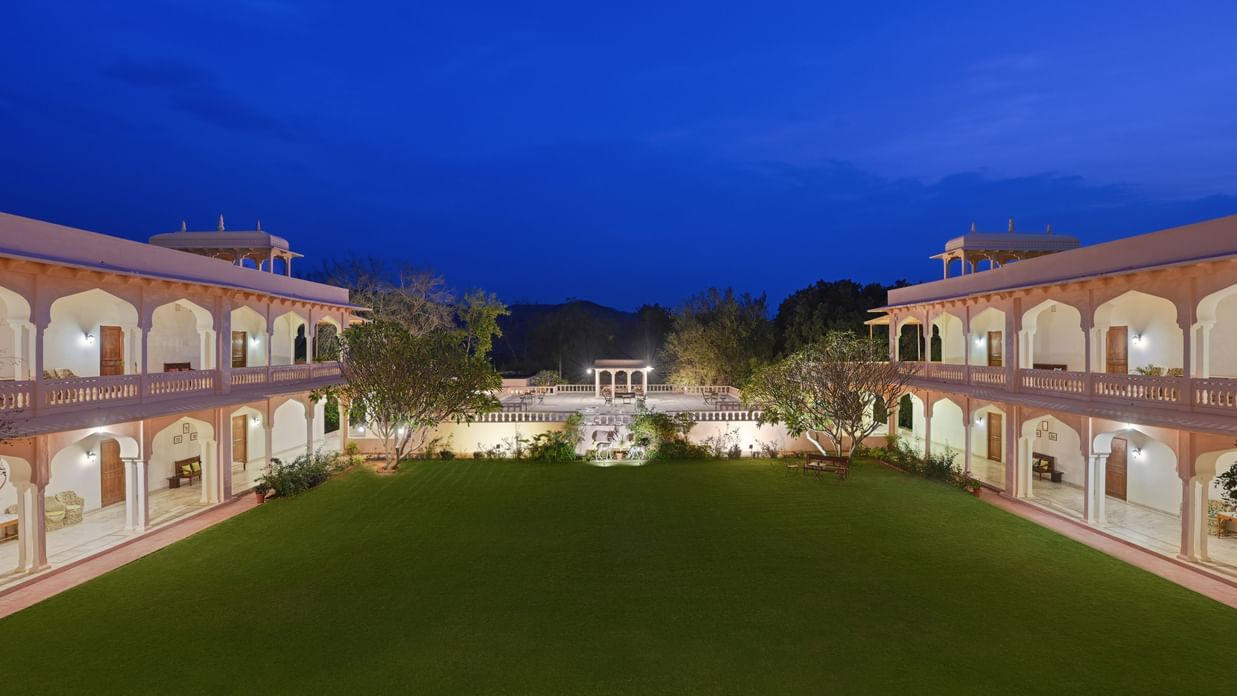 Facade of Hotel Dev Vilas, Ranthambore illuminated during late evening with an expansive lawn with silhouettes of trees in the background
