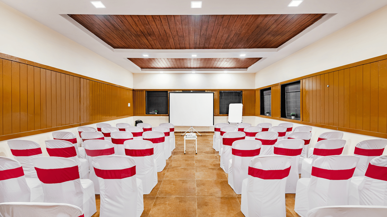Nature Trails Kudalika conference hall rear view with white and red theatre chairs, central projector stand, projection screen and dark wood coffered ceiling.