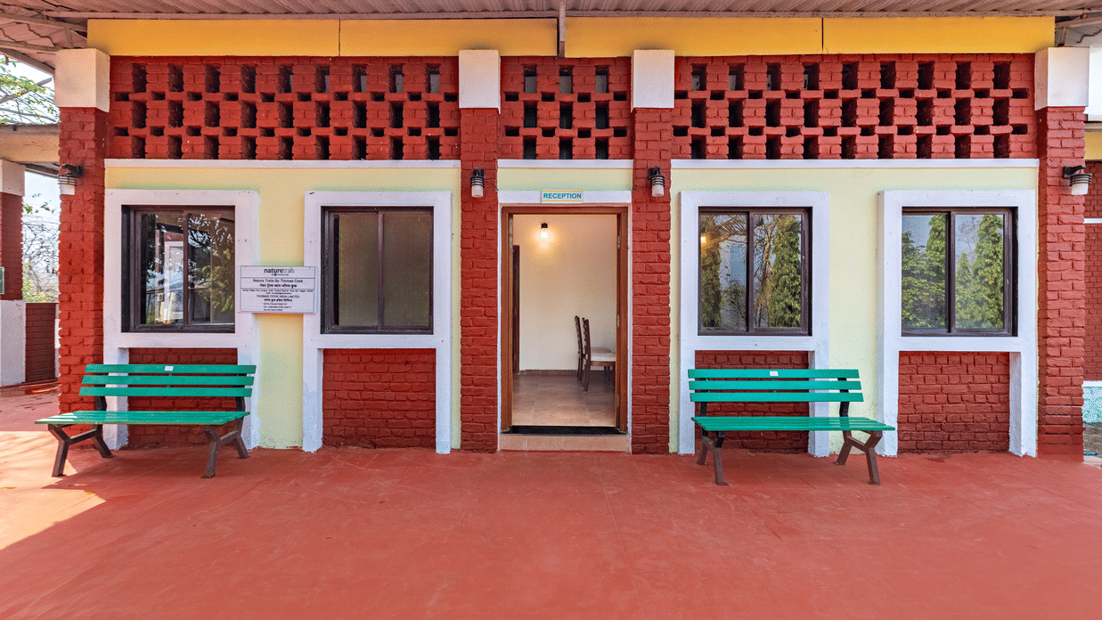 Nature Trails Kudalika reception building front view with red laterite brick walls, yellow band, latticed brick top, two green benches and open entrance door.