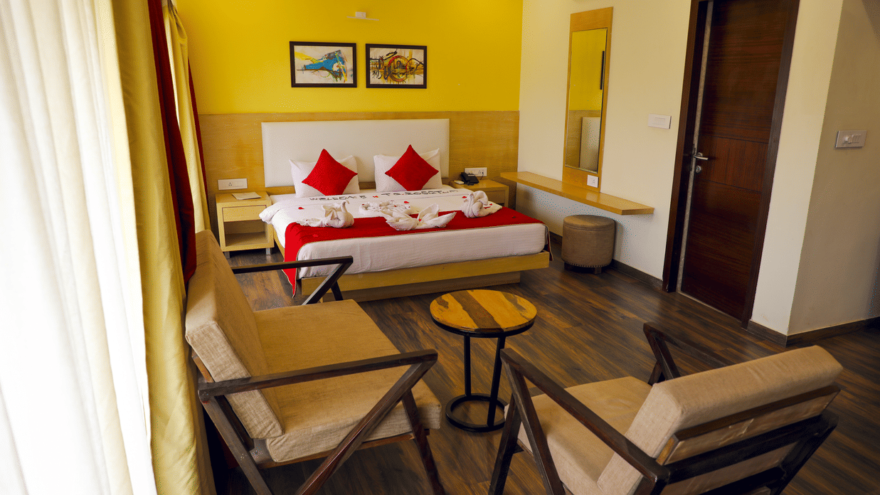 A hotel room with a queen bed, white and red linens, and a bright yellow accent wall. The bed is flanked by light wood nightstands.
