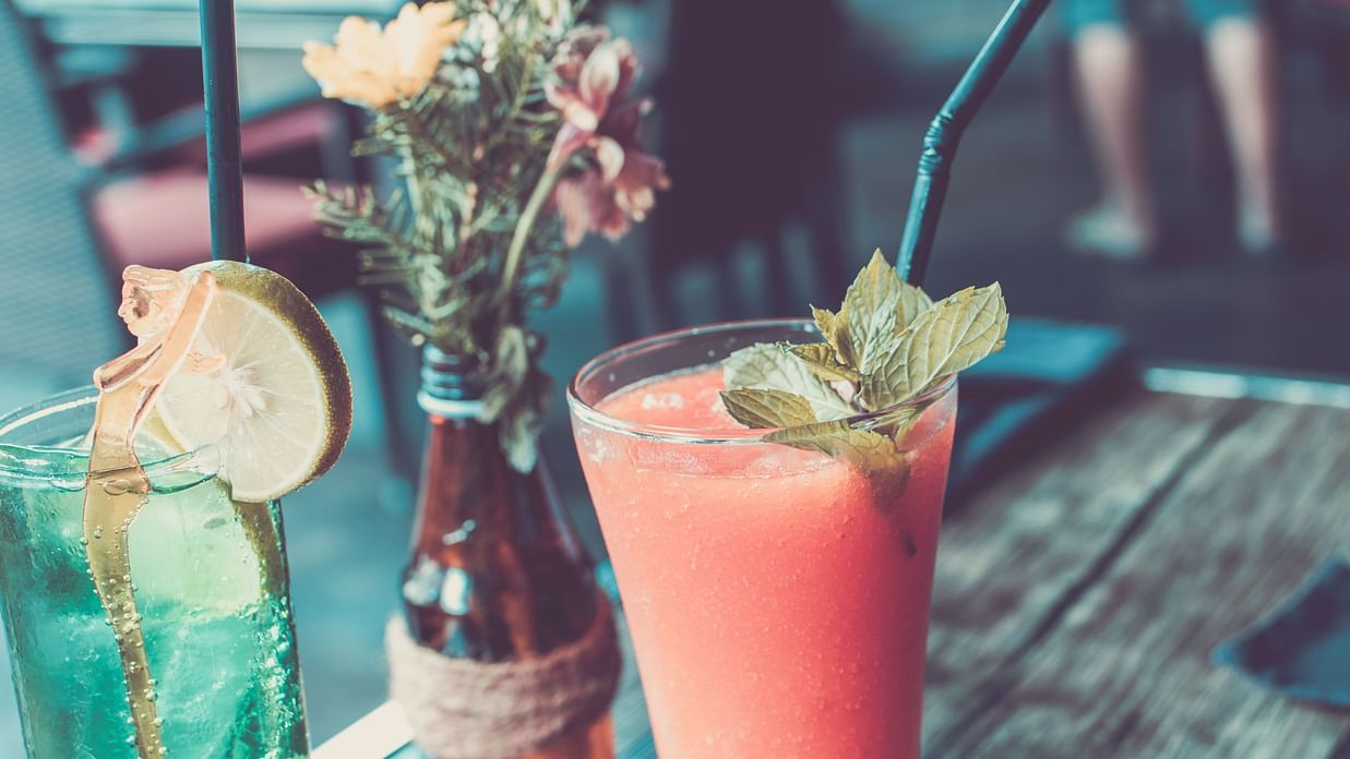 Two refreshing cocktails served on a wooden table, garnished with mint and lemon, next to a small flower arrangement in a bottle.