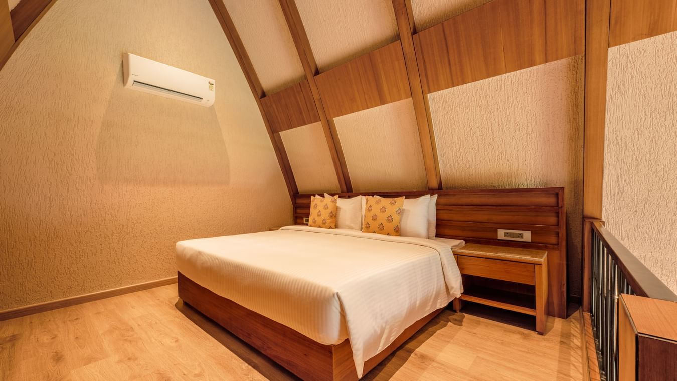 Plush double bed seen beside a wall with a wall-mounted air-conditioning, bedside tables, wooden flooring, and A-shaped high ceiling.