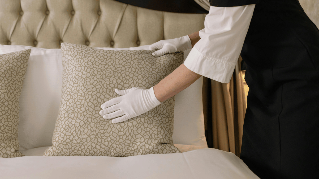 Housekeeper in an uniform and gloves smoothing a decorative pillow on a neatly made hotel bed.