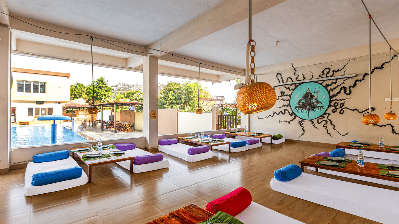 Spacious open-air dining hall with low wooden tables and floor cushions overlooking a swimming pool - Nature Trails Ashoka Resort Hampi