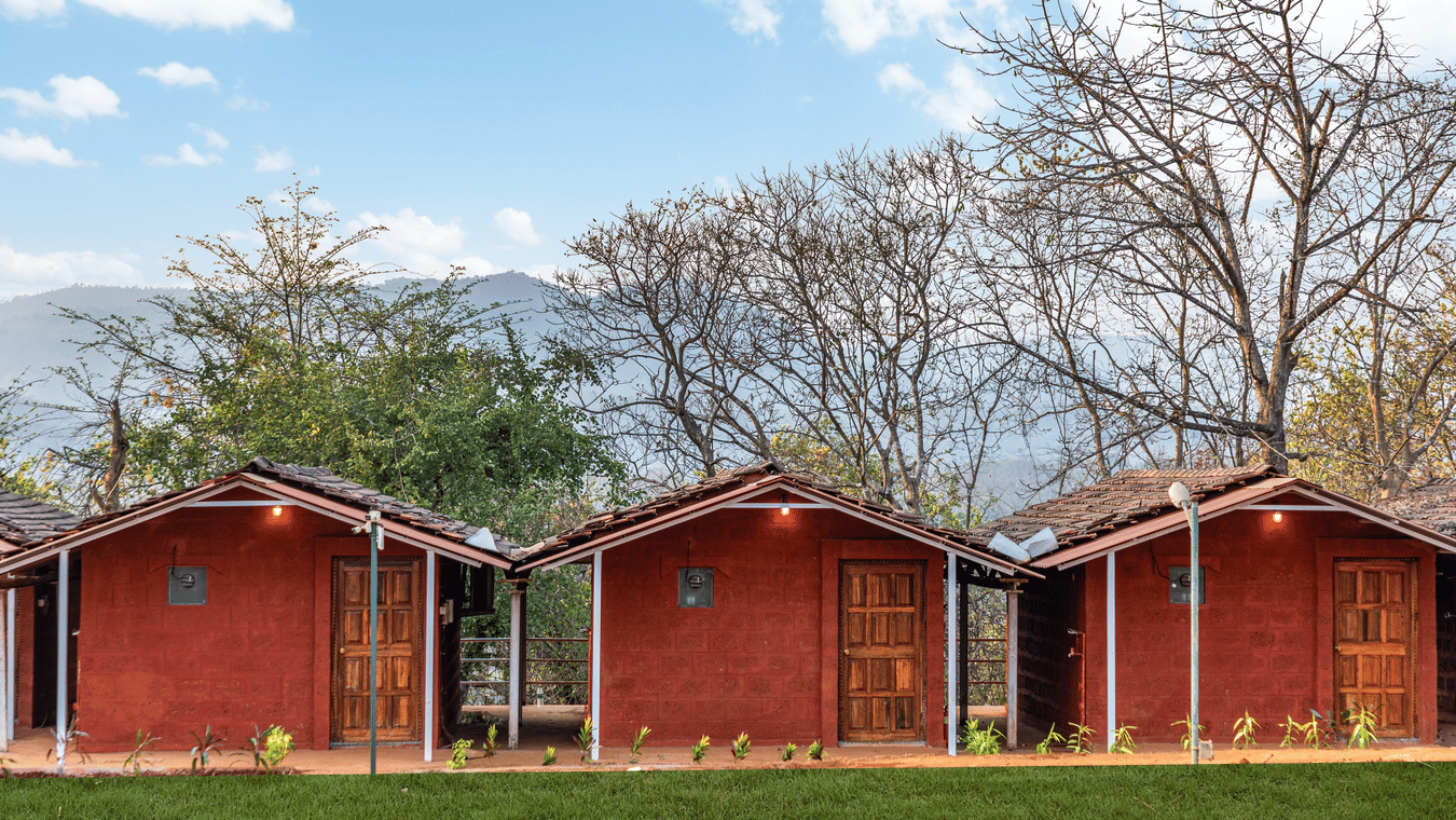 Nature Trails Kudalika three red laterite cottages with wooden panel doors, tiled roofs, wall lights and small verandahs, set against mountain and tree backdrop.