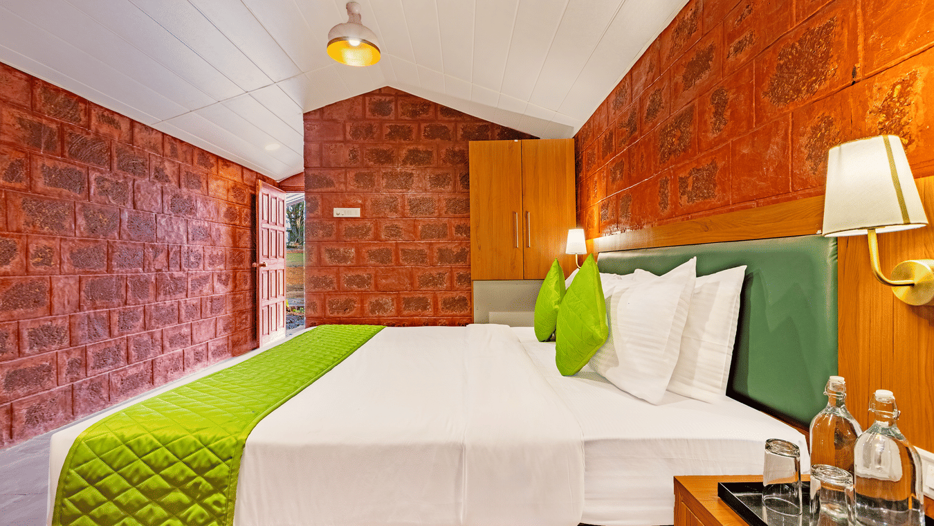 Nature Trails Kudalika premium heritage room with double bed, green bedding, red laterite stone walls, wooden wardrobe, open entrance door and green outdoor view.