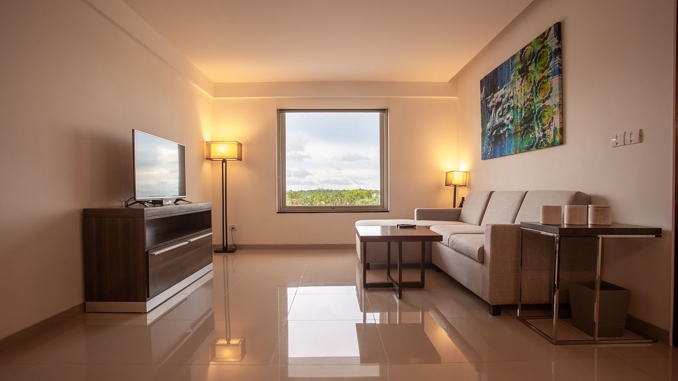 A living area with a comfortable sofa, a TV unit, and a table, featuring a portrait on the wall and a window overlooking the outside at The Residency Towers. Rameswaram