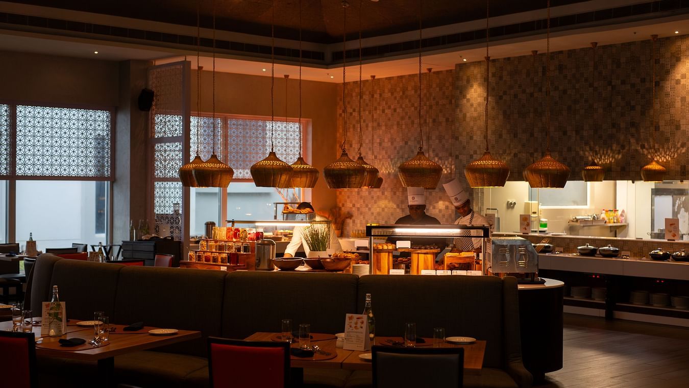  A modern restaurant interior with a buffet counter, chefs cooking, and tables set for dining. The restaurant has warm lighting and decorative hanging lamps at THE RESIDENCY TOWERS RAMESWARAM