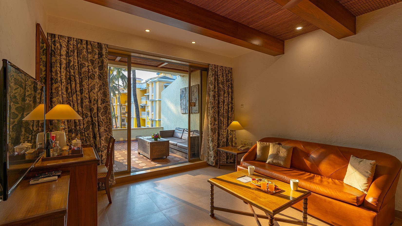 A living room that includes a sofa, coffee table, tea and coffee maker, a TV, and an outdoor balcony with seating arrangements.
