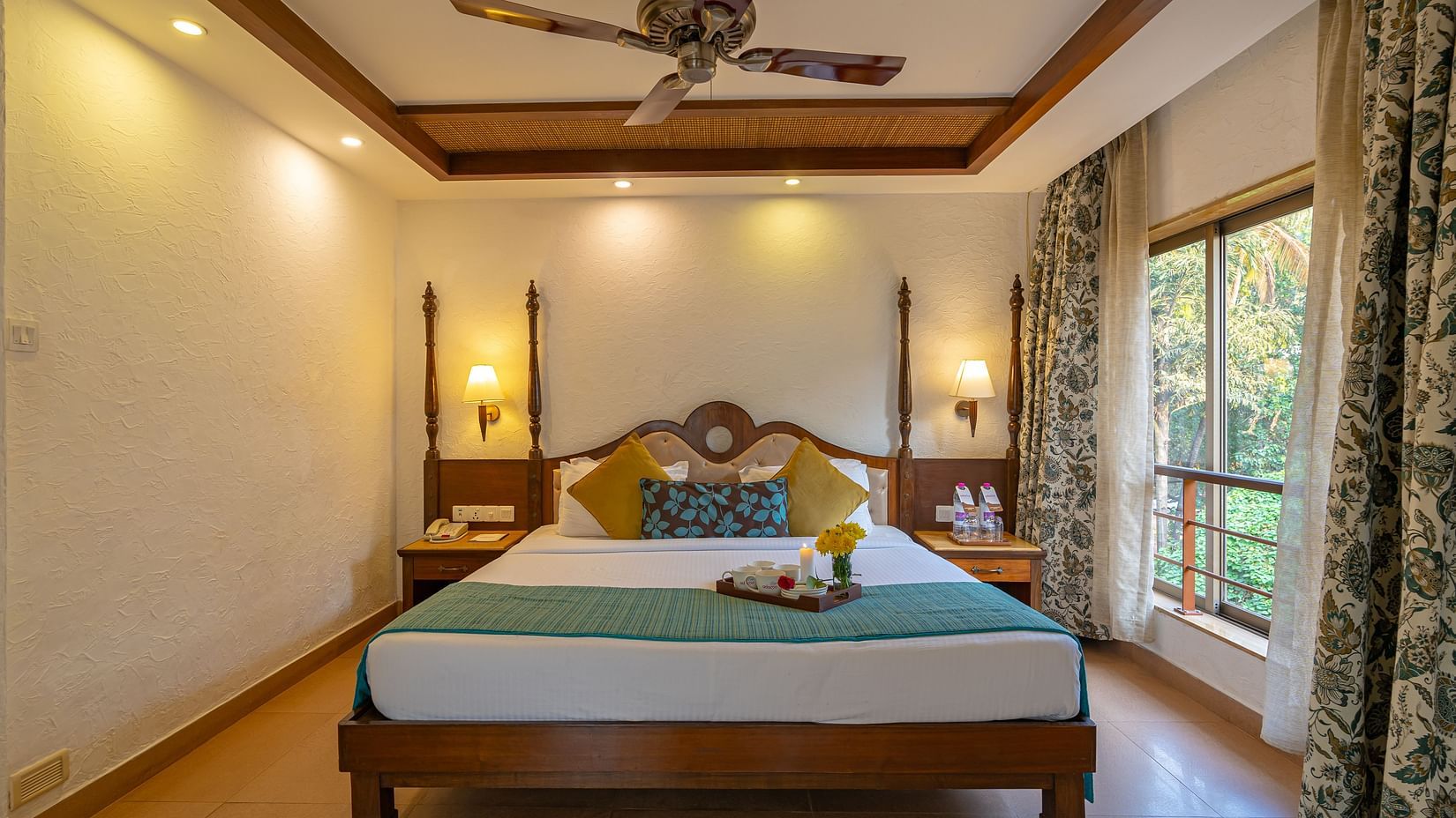 An image of a hotel room featuring a beautifully decorated bed with a tray of cups, plates, and flowers, complemented by a large window, a fan, and table lamps.
