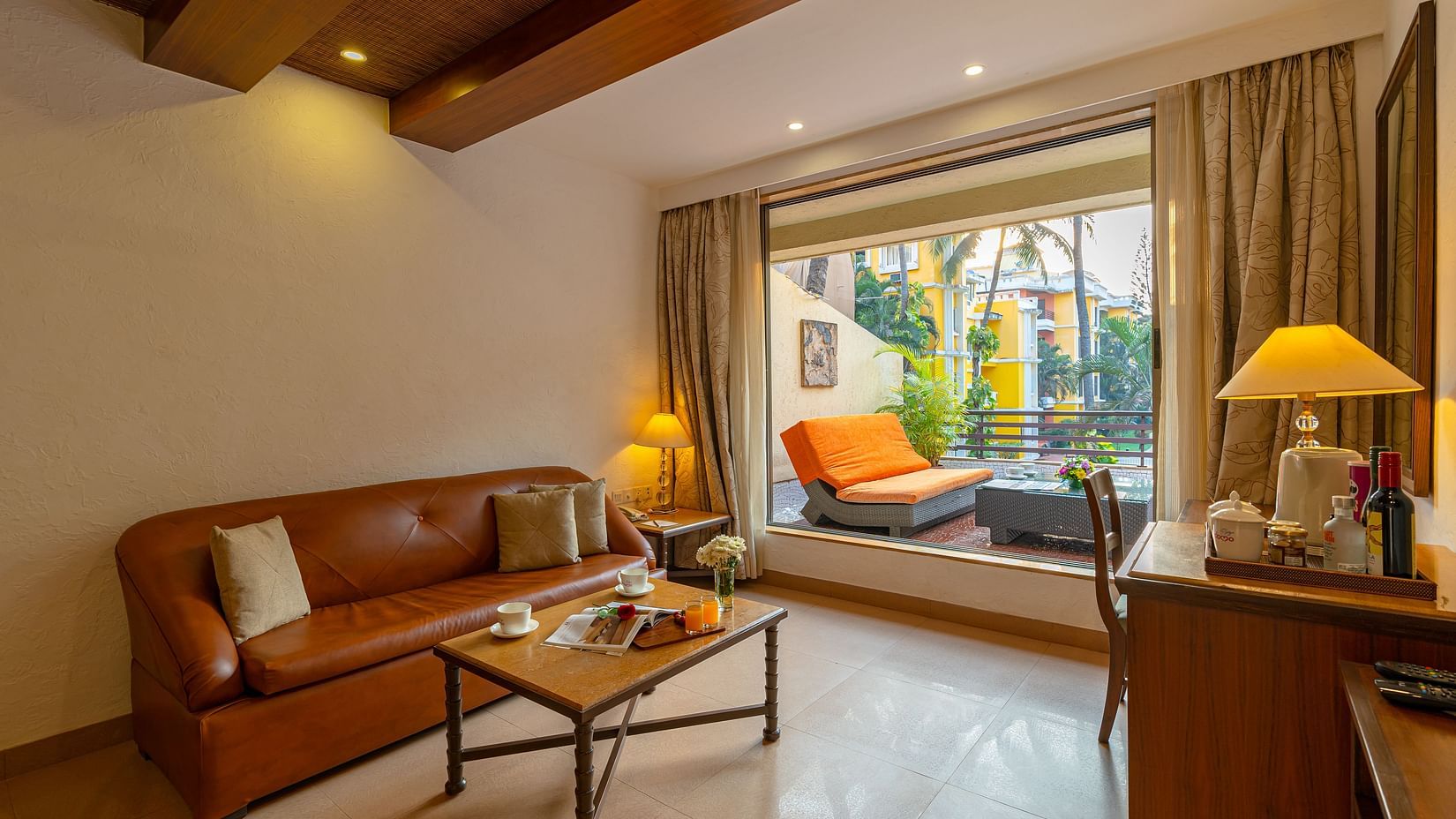 An image of a living room with a brown sofa, coffee table, table lamps, a tea and coffee maker, and an attached balcony with seating.