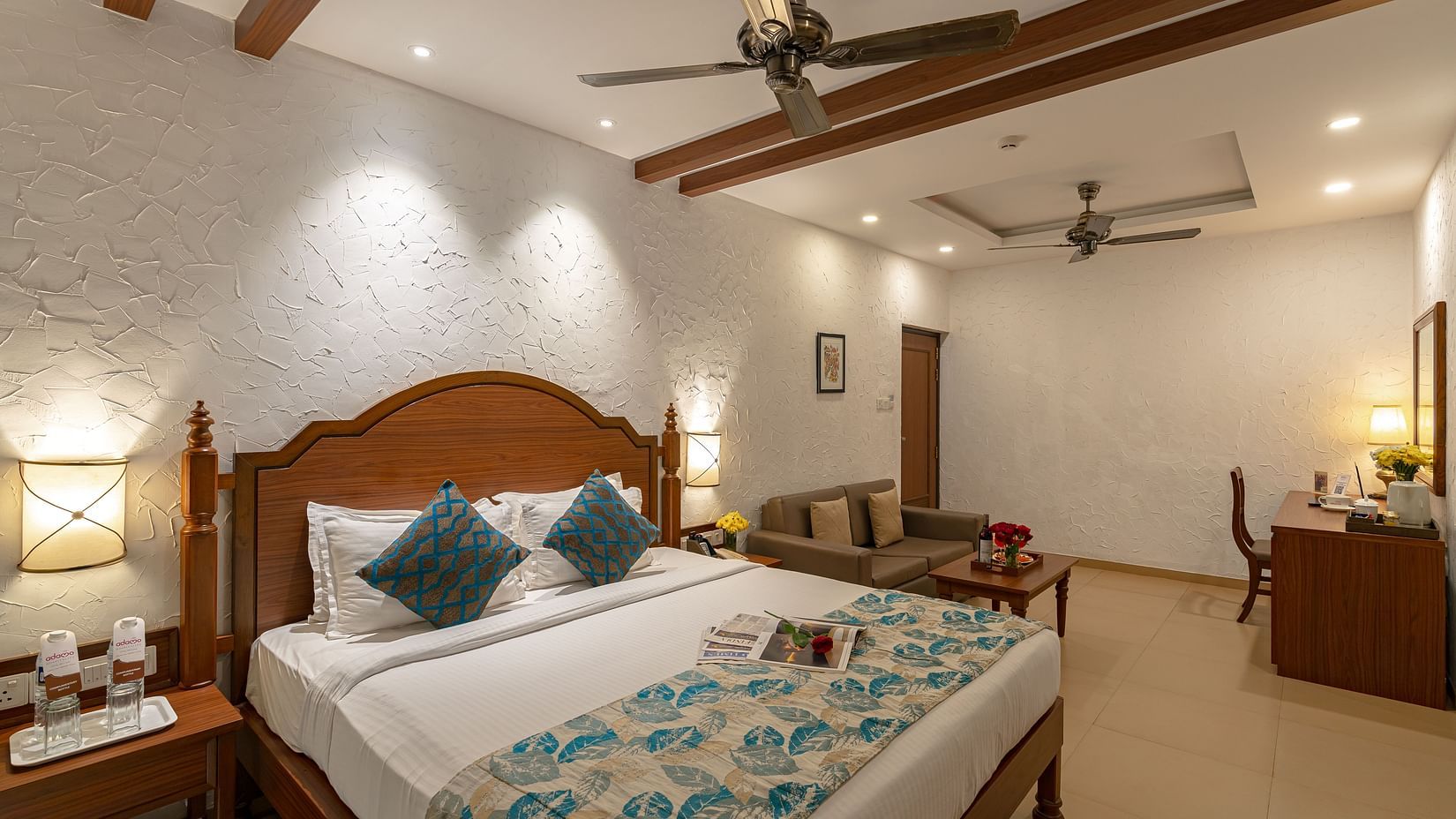 A hotel room featuring a neatly made and decorated bed, two wall-mounted lamps, a sofa, a side drawer, and a vanity.