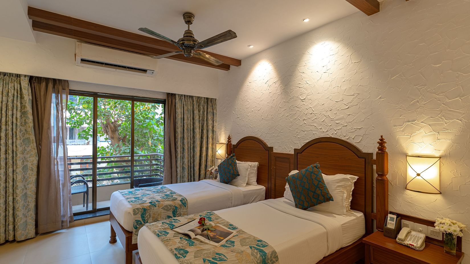 An image of a hotel room with twin beds dressed in white sheets, a cosy throw, wall-mounted lamps on each side, and an attached balcony.