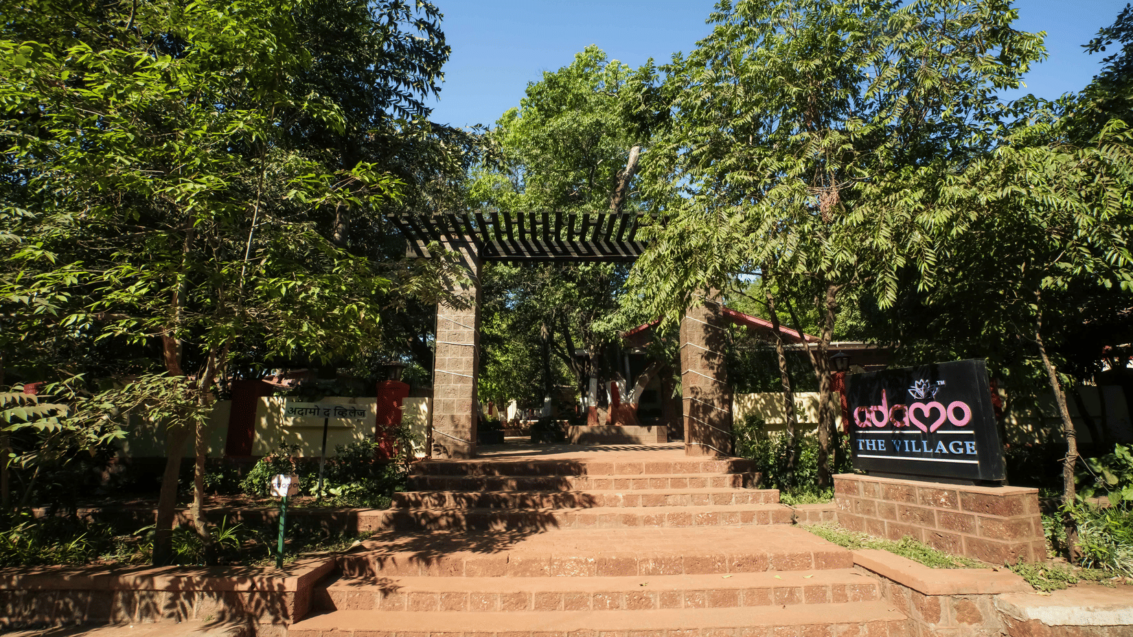 Outdoor view of wide stepped entrance path leading to Adamo The Village, surrounded by tall tress and greenery.