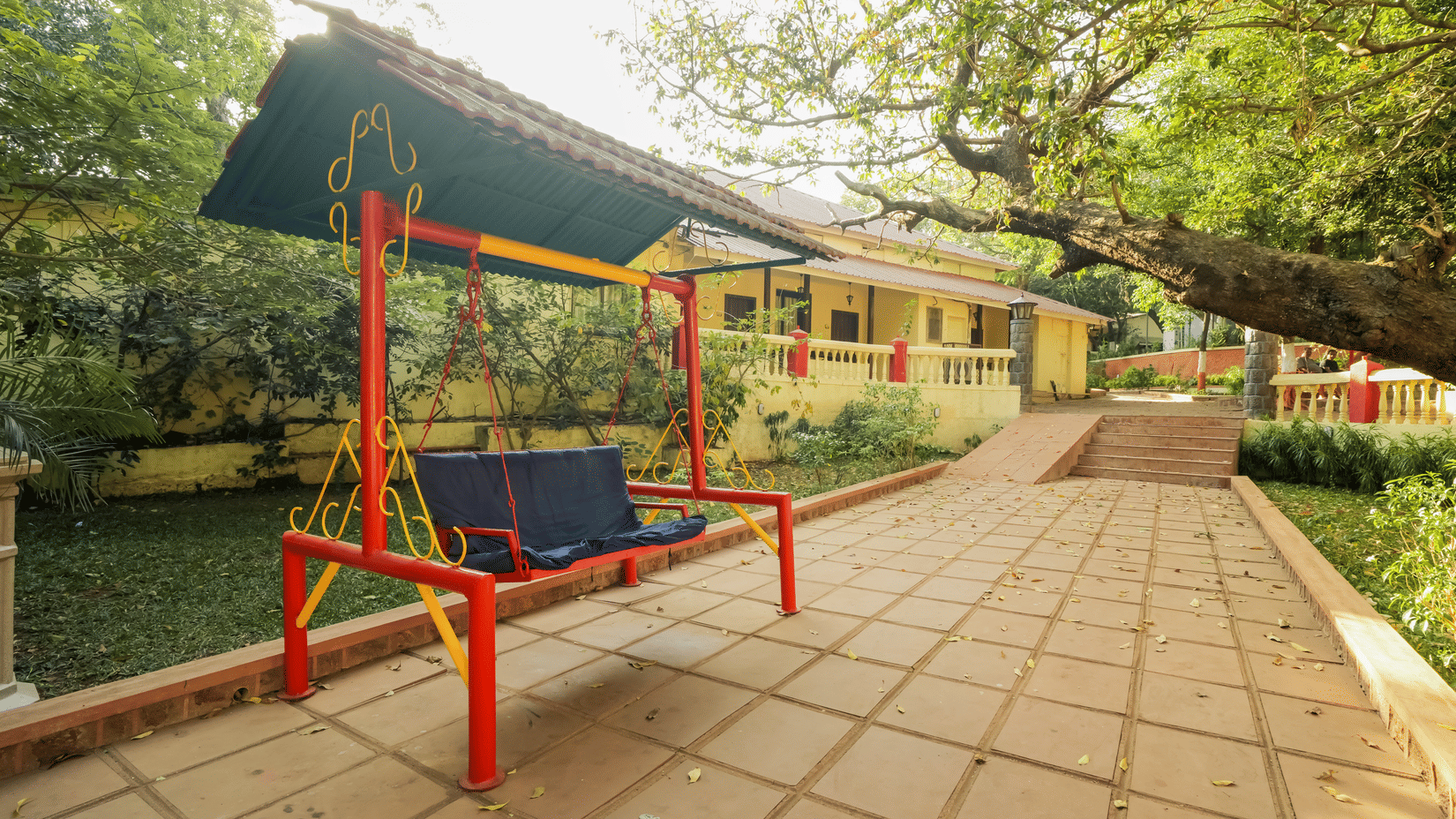An outdoor paved pathway featuring a red-framed covered swing bench surrounded by green trees and resort buildings.