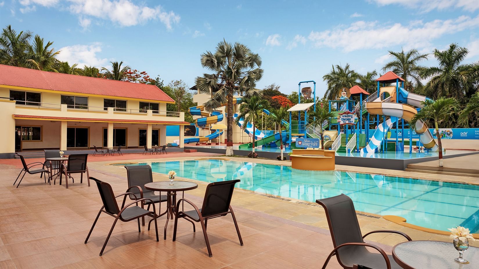 A swimming pool at DGV Resort by Treat, Silvassa with colourful slides, splash structures, a swimming pool, poolside seating, palm trees, and a building with red-tiled roof.