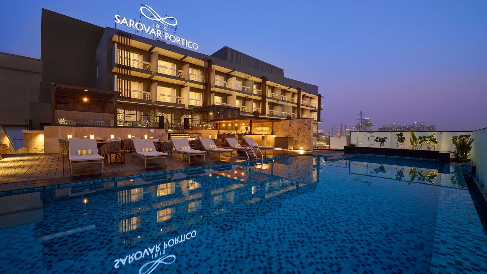 A modern hotel building is illuminated at dusk, featuring a rooftop swimming pool reflecting the hotel's signage and nearby lounge chairs. - Iris Sarovar Portico, Sector 85, Gurugram