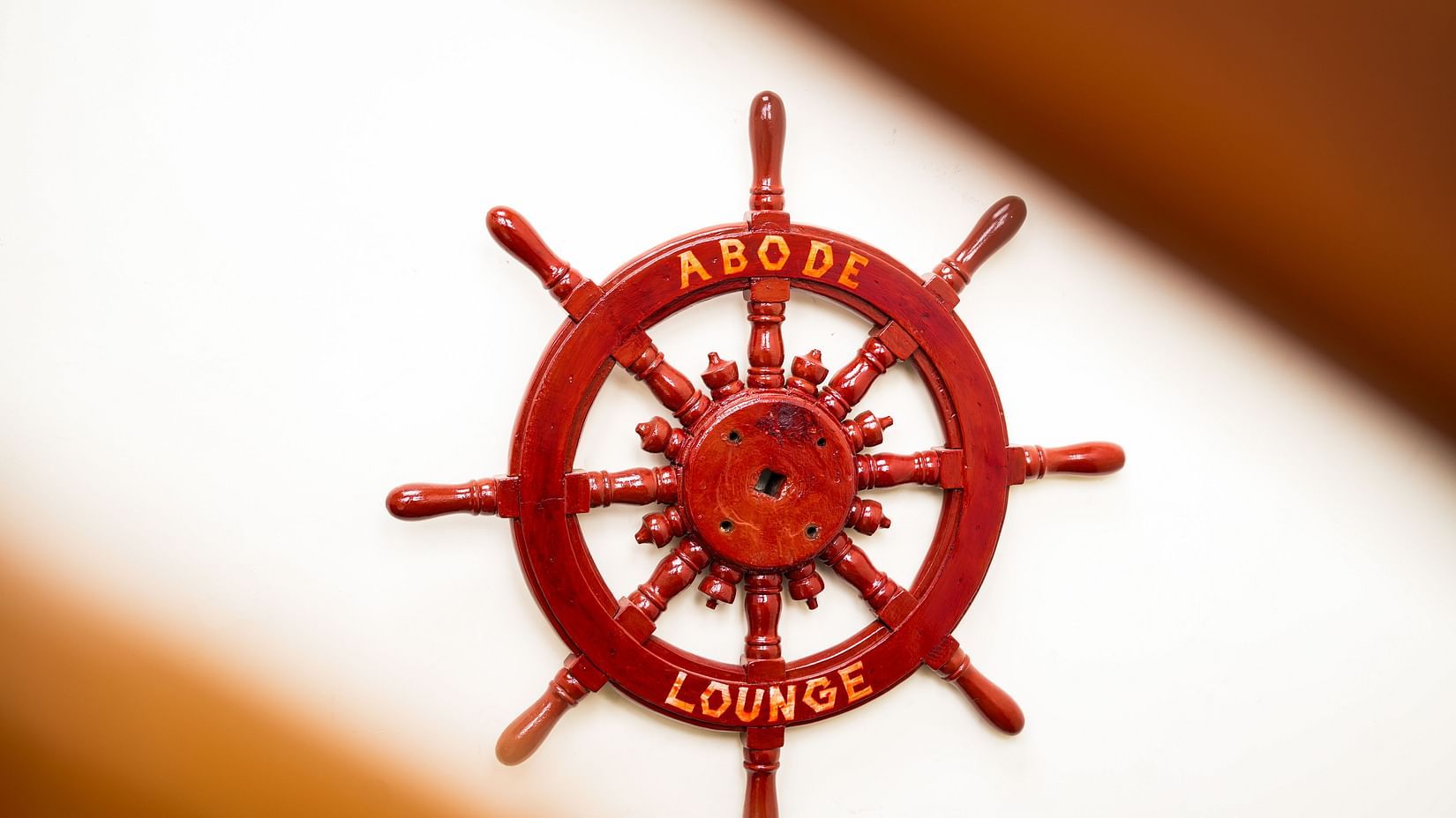 A classic wooden ship's wheel painted in deep red, featuring the words ABODE LOUNGE in yellow, mounted on a white wall - MGM Beach Resorts
