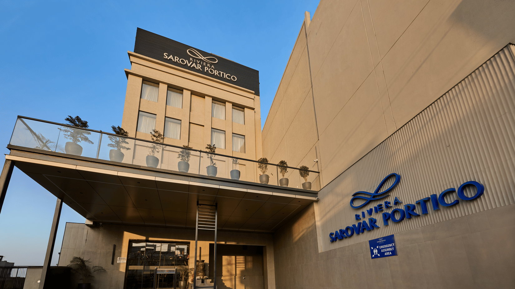 Exterior view of Riviera Sarovar Portico showcasing the main entrance, modern façade, and hotel signage, captured during daylight with a clean architectural perspective.