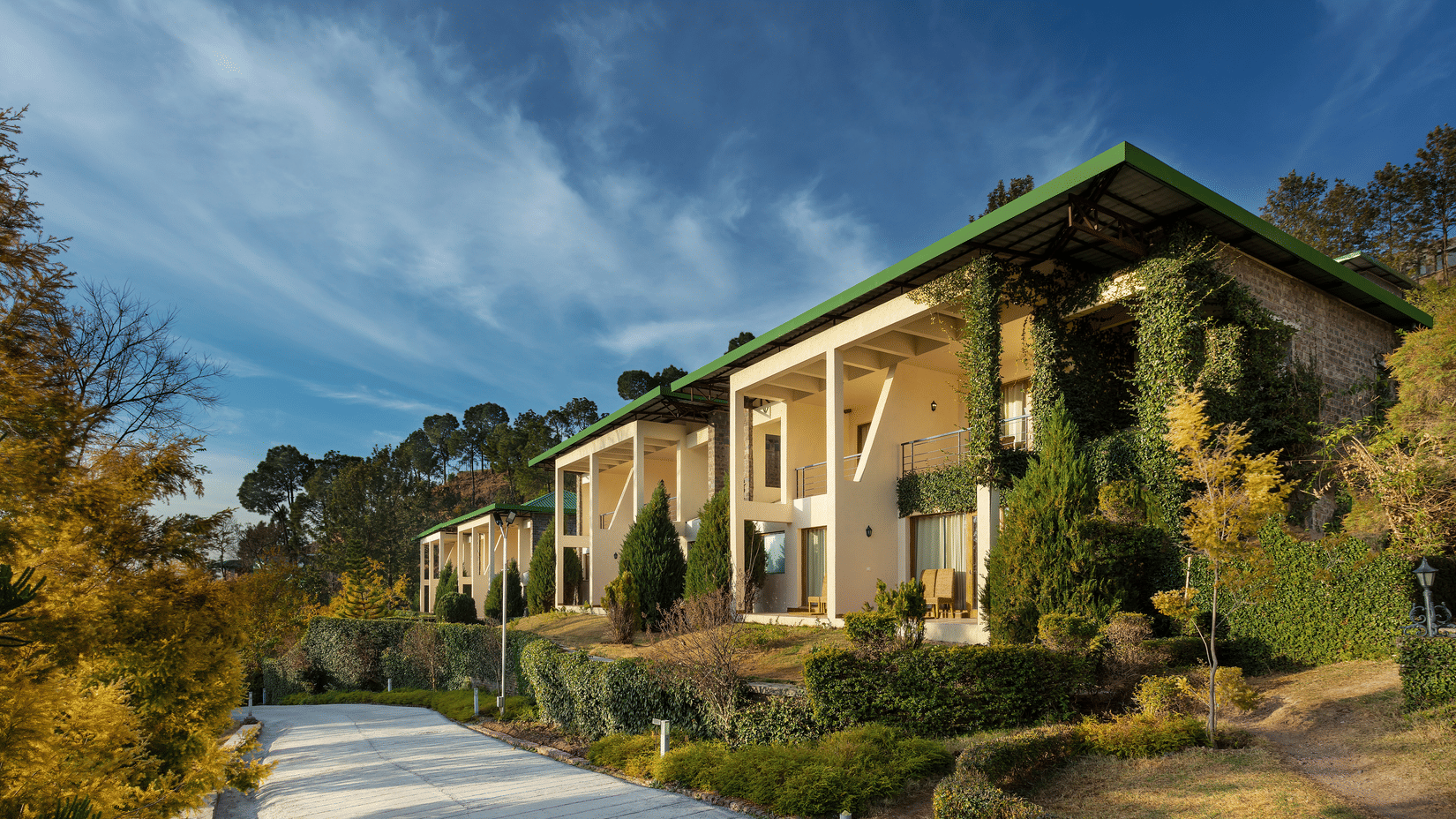 Facade of Suryavilas Luxury Resort and Spa in Solan with a paved driveway and green climbing plants on its exterior walls, under a blue sky with clouds