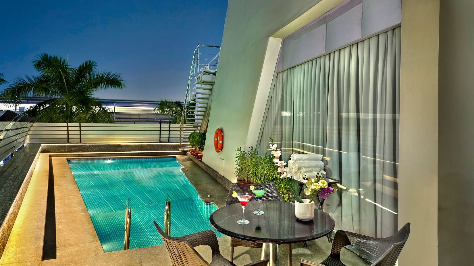 A dimly lit outdoor patio featuring a small, brightly lit pool, comfortable lounge chairs, and a round table with floral arrangements- The Sonnet, Kolkata