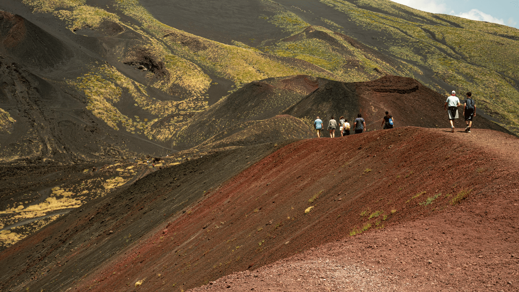 A group of people walking along the ridge of a reddish-brown hilly landscape with sparse vegetation.