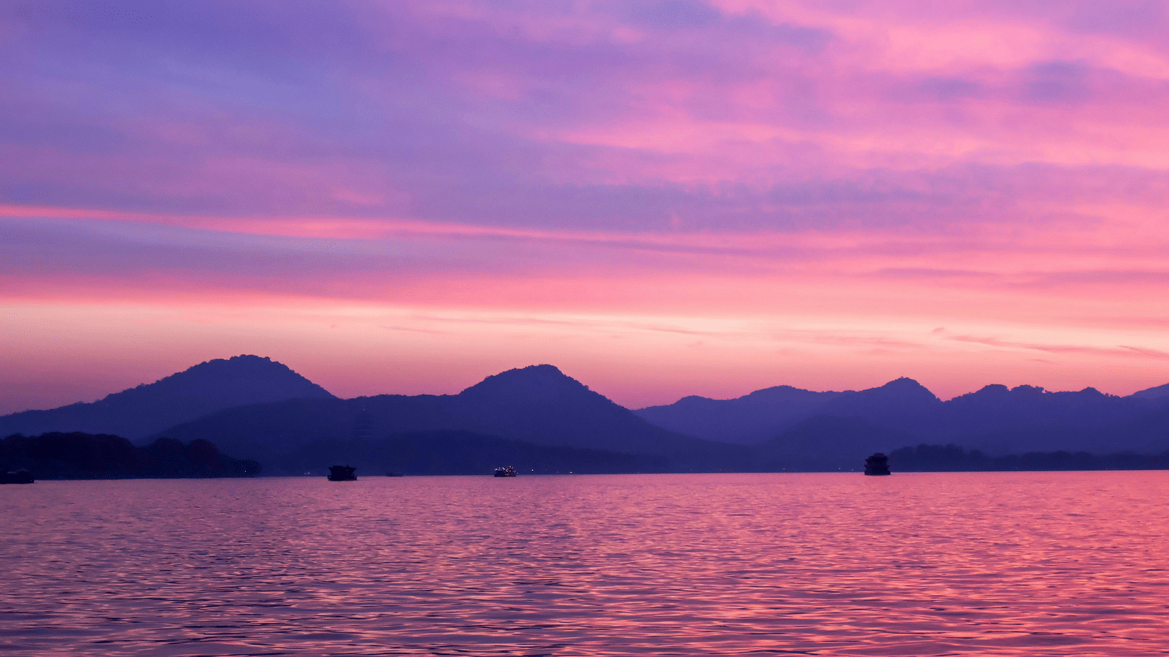 Pink and purple sunset over Fateh Sagar Lake with silhouetted mountains and small boats drifting across reflective water surface