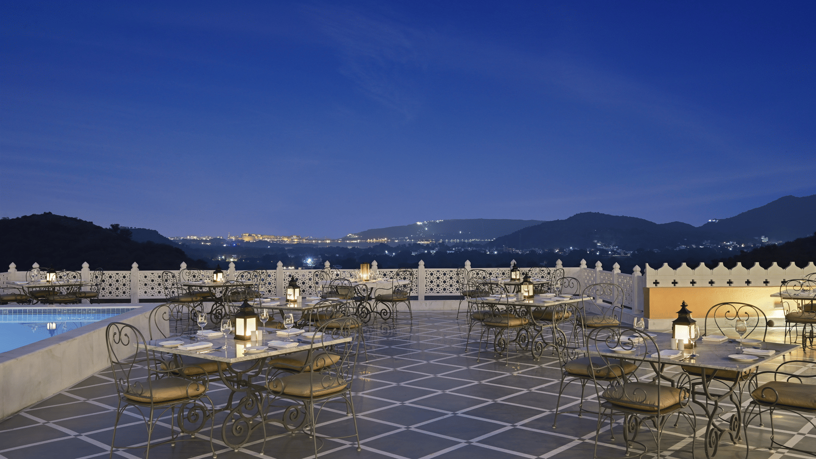 Terrace next to the swimming pool with seating arrangement for fine dining in Udaipur at Fateh Vilas and views of the mountains in the twilight sky