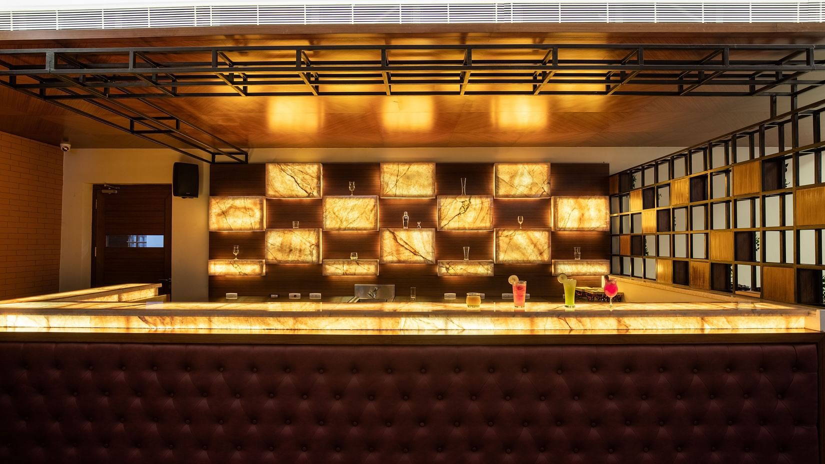 A dimly lit bar area with a cushioned counter, illuminated shelves, and abstract wall art - Jenneys Residency, Coimbatore