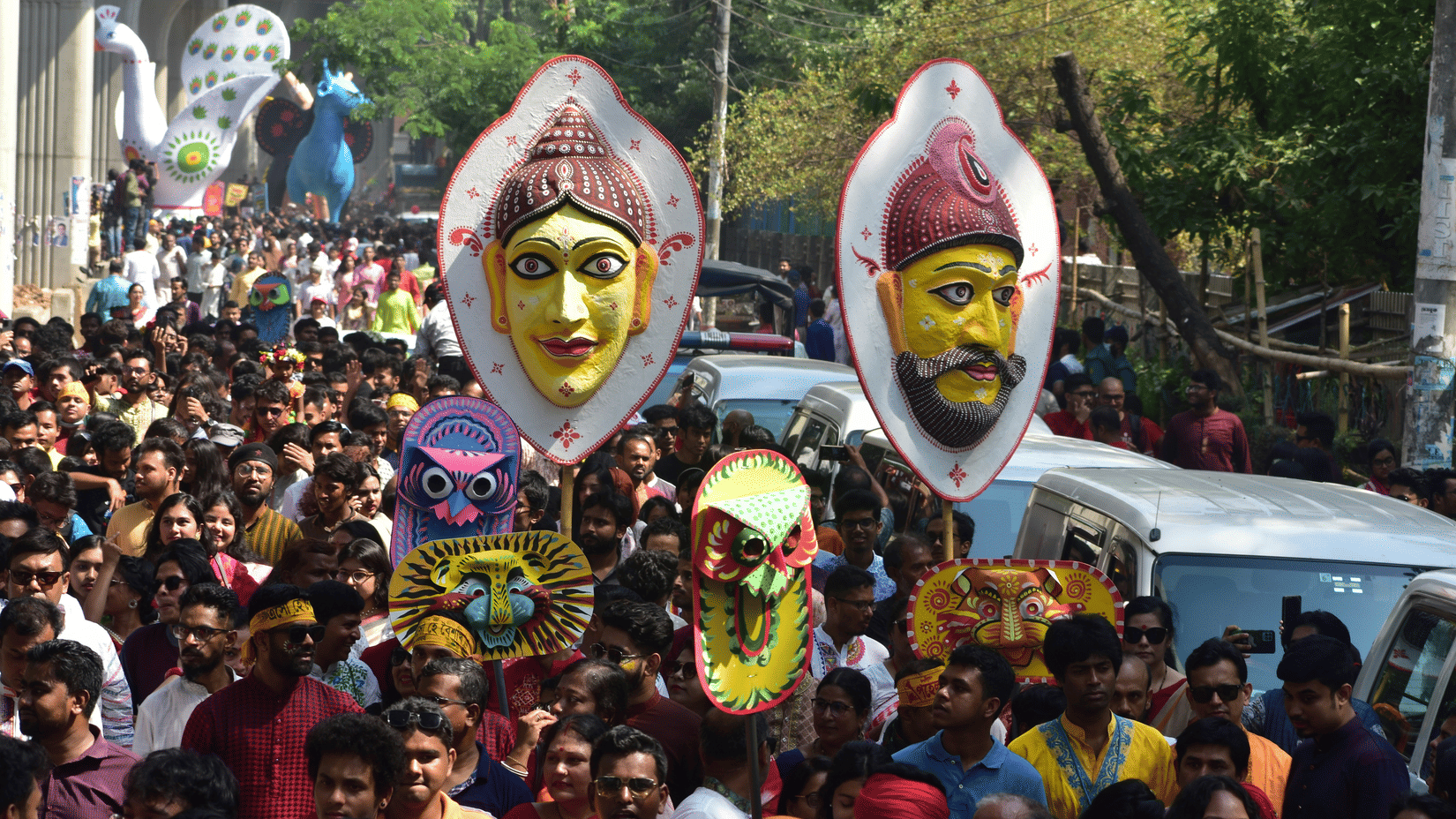 A procession on a road with many people carrying busts on a pole next to parked cars during the Bengali New Year celebration in Kolkata.