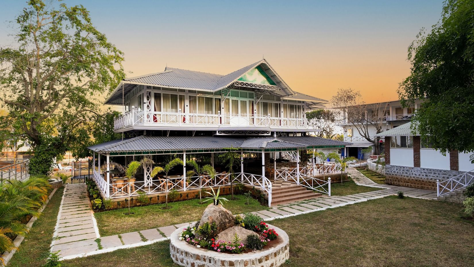 The facade of the Virundhu Restaurant with a two tier building with trees and a small rock garden in the front area.