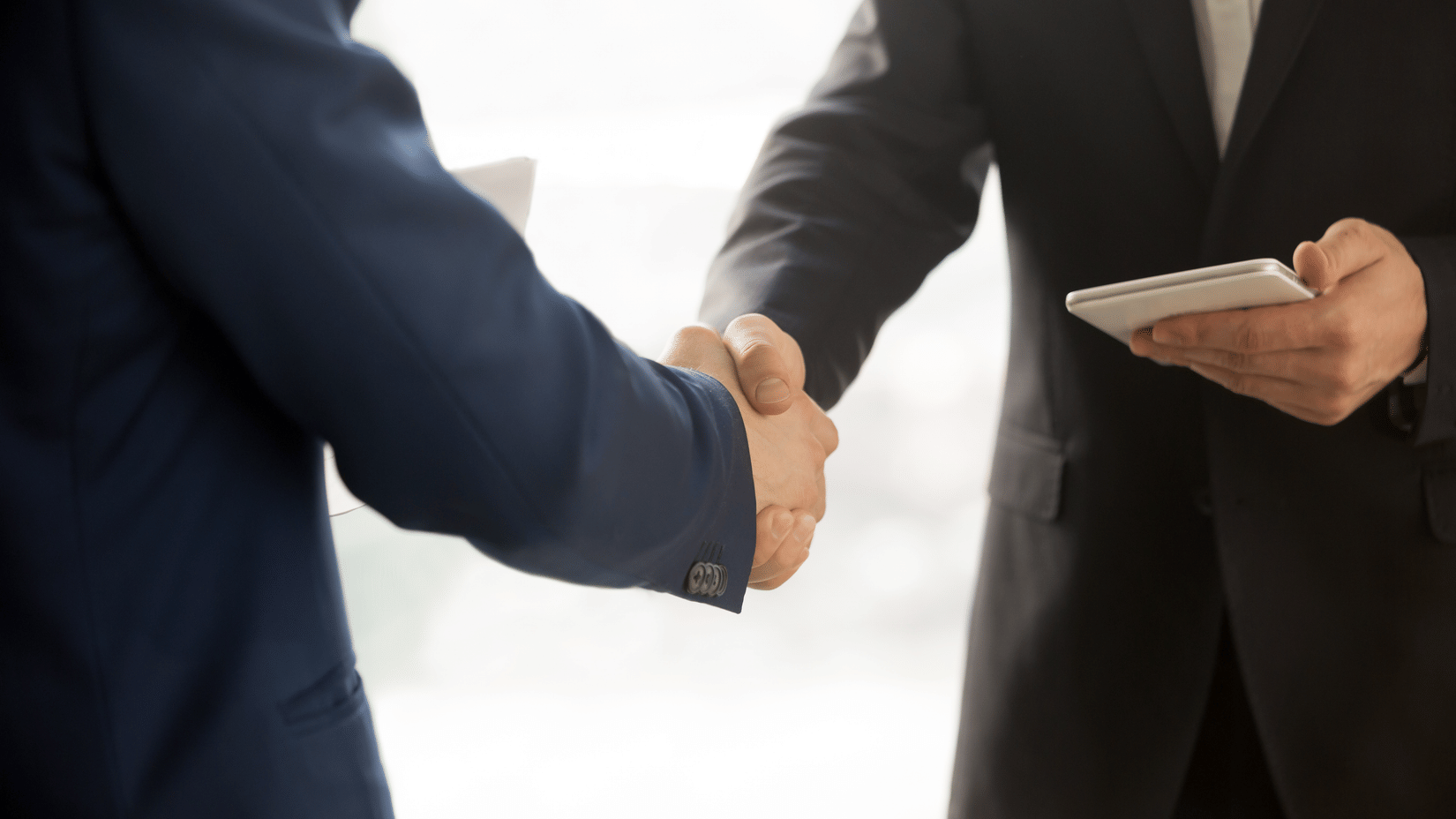 Two people in suits shake hands, symbolizing a professional agreement or partnership. One holds a tablet, suggesting a business context.