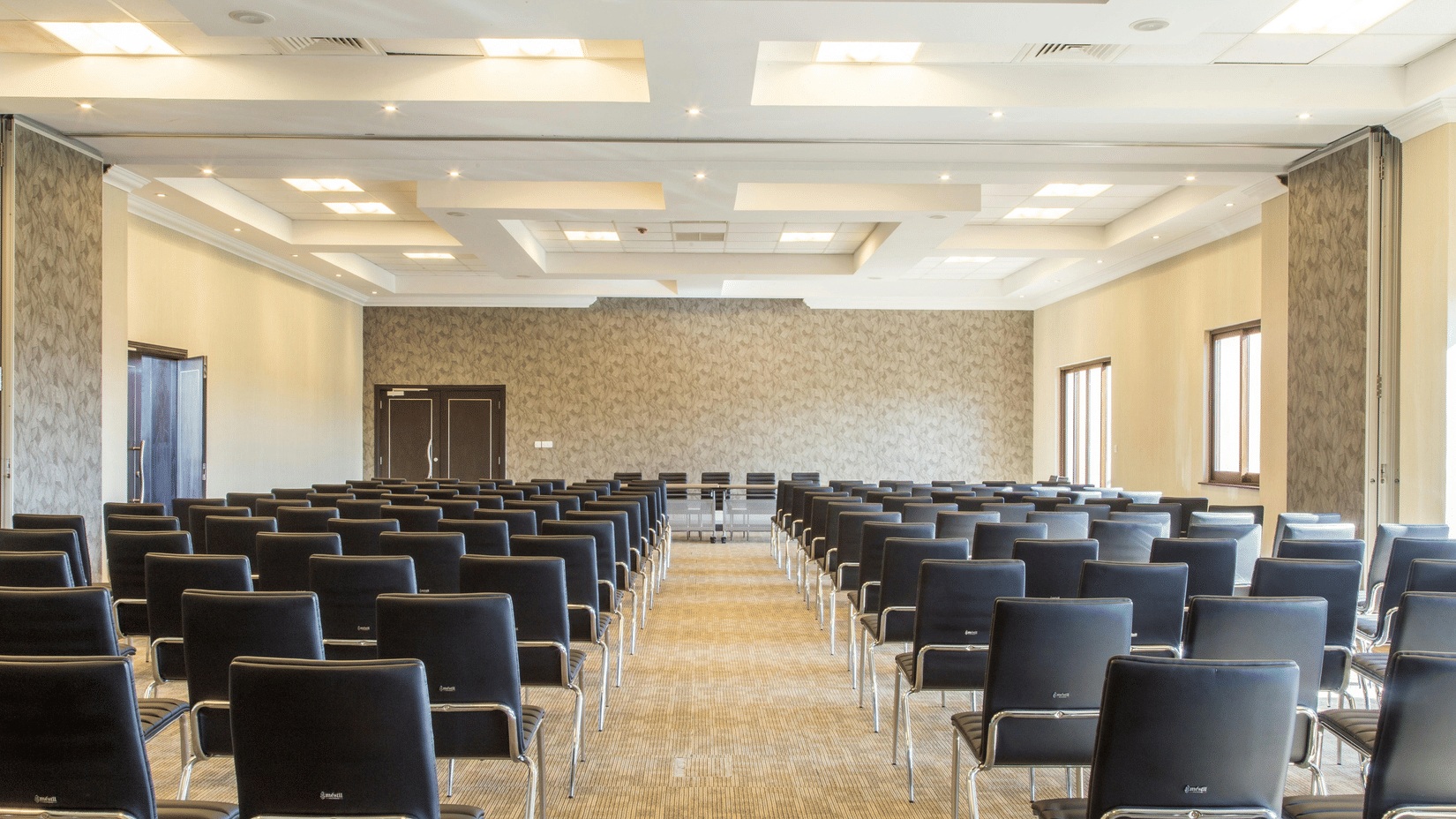 A conference room with rows of black chairs neatly arranged facing a long table at the front. The room has beige walls, patterned wallpaper at the far end, large windows on the right side, and a ceiling with recessed lighting