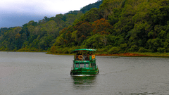 A green boat cruises across a calm river framed by lush, forested hills and an overcast sky.