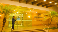 A security guard stands next to a large glass door entrance at Ambar Sarovar Portico, Gandhidham featuring steps, overhead beams, and palm trees.