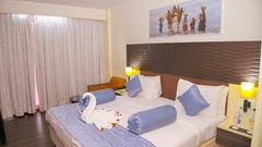 A hotel guestroom at Ambar Sarovar Portico, Gandhidham is furnished with a king size bed, bedside lamps, and a decorative painting above the headboard.