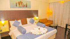 A hotel bedroom at Ambar Sarovar Portico, Gandhidham shows a large king size bed decorated with folded towels, a sofa, and a wall painting.