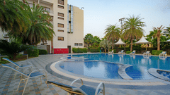 View of the outdoor swimming pool area with palm trees and the main hotel building visible in the background at Arawali Ananta Elite, Jaipur.
