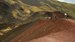 A group of people walking along the ridge of a reddish-brown hilly landscape with sparse vegetation.