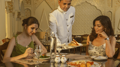 Two women enjoy a gourmet meal as a server in a white uniform presents a dish in a luxurious restaurant with intricate decor.