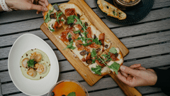 Two people sharing pizza on a wooden platter with side dishes around